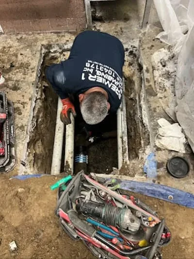 Plumber working in excavated sewer line pit with tools and equipment visible