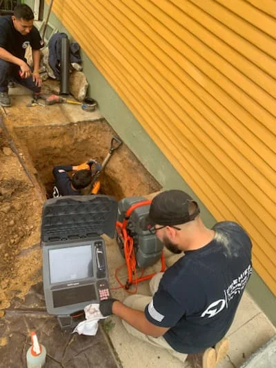 Plumbing technician operating sewer camera equipment in excavated pit with yellow corrugated siding visible