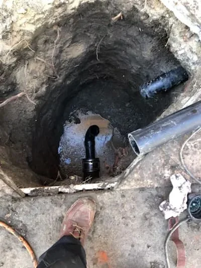 Excavated sewer line pit showing black pipe with water accumulation and new blue PVC connection
